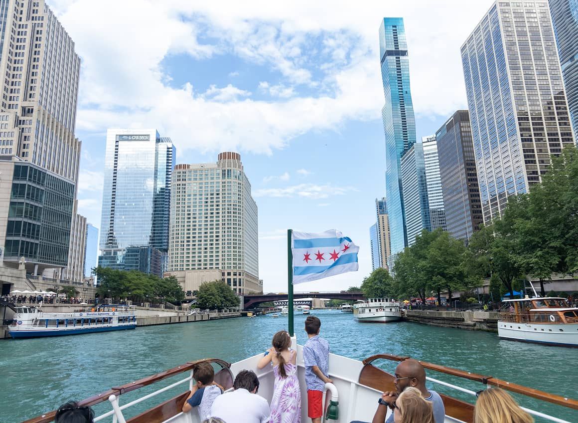 Photo of tourists on a boat in the Chicago River with the Chicago flag at the bow. 